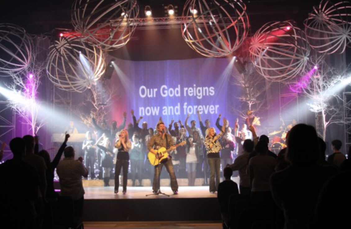 A trio singing in front of a crowd with a choir in the background